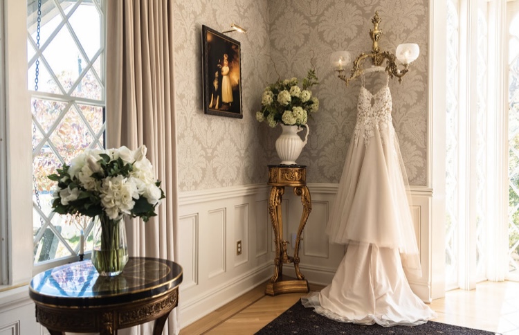 Cream-colored corner with side tables, vases with flowers, and a hanging wedding dress