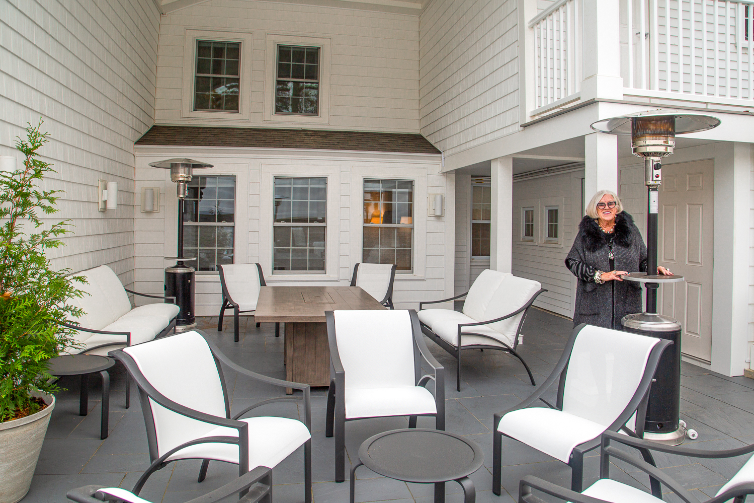 Gail stands on the patio with sleek patio furniture in front of a historical building with white siding.