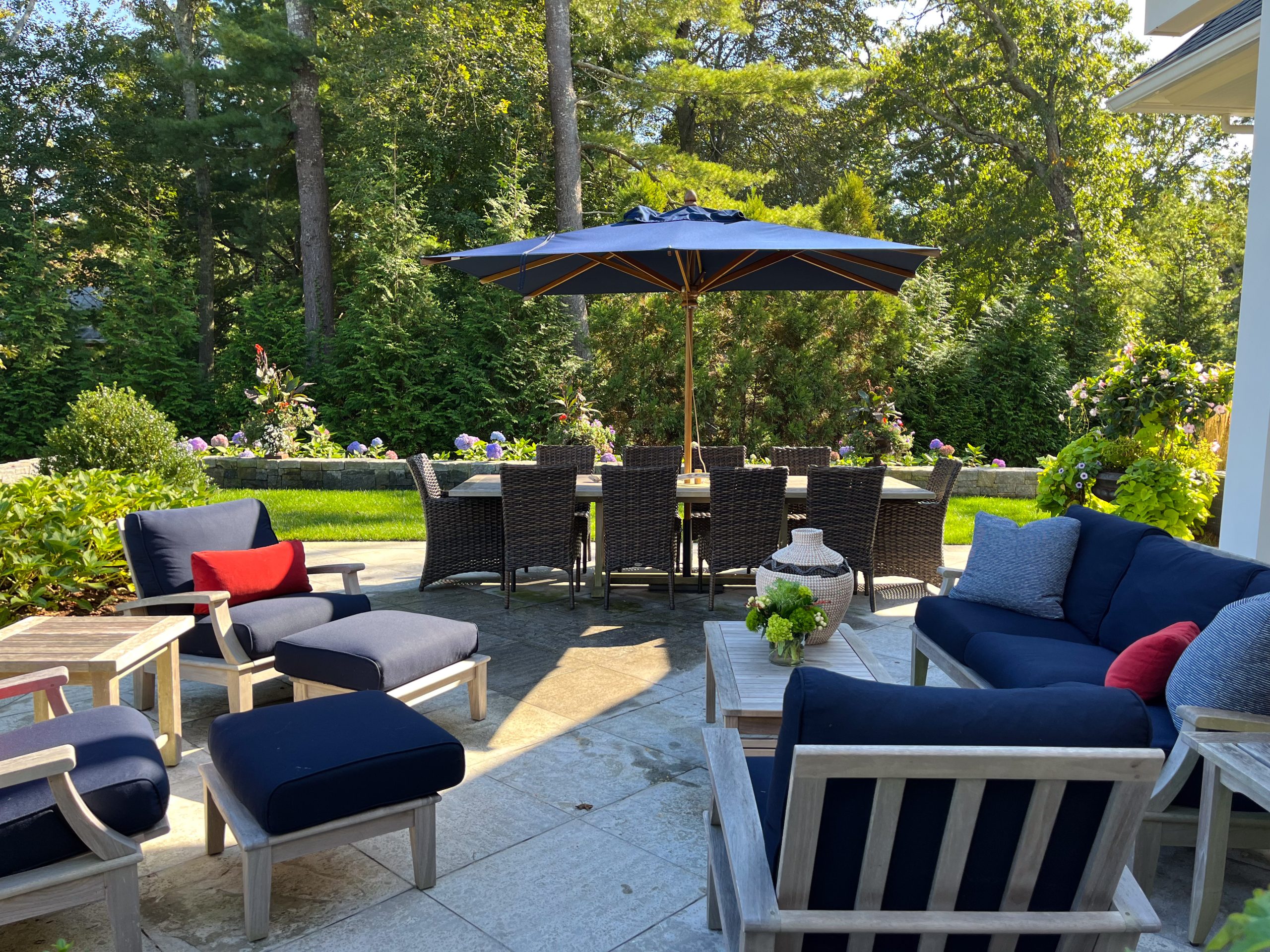 A beautiful yard with green trees and a patio set with navy blue cushions and red accent pillows.