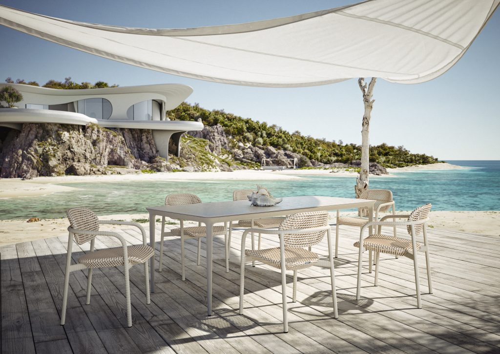 An outdoor dining table and chairs on a wood deck at the beach with a white fabric shade overhead, and a luxurious modern mansion in the background