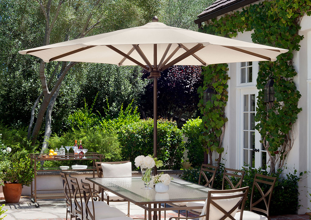 An outdoor dining table in a beautifully landscaped backyard with a bronze and white outdoor shade umbrella over it