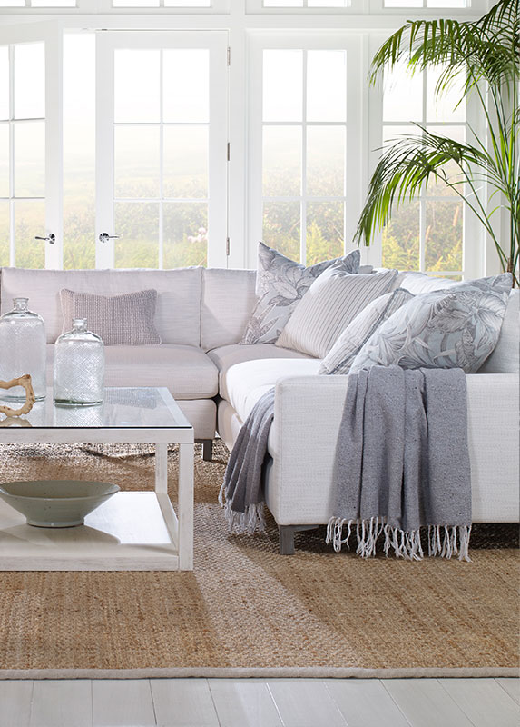 A coastal themed living room with large windows, a cream sofa, and a natural woven grass rug